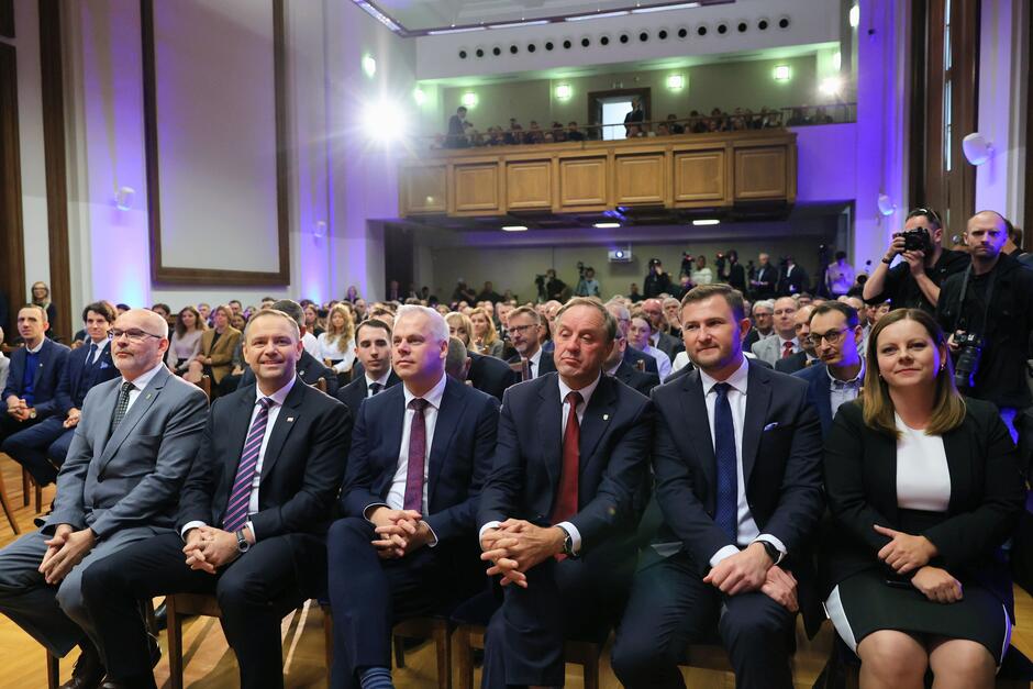 Na zdjęciu widać elegancko ubranych uczestników wydarzenia siedzących w pierwszym rzędzie w dużej sali, pełnej ludzi. W tle znajdują się widzowie na balkonie oraz fotografowie dokumentujący wydarzenie. 