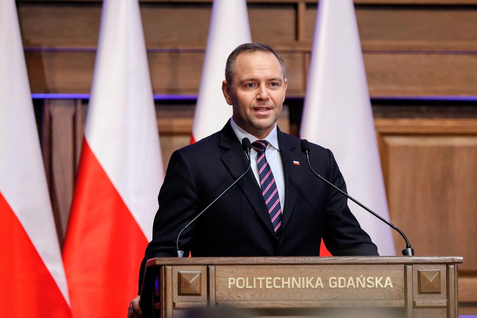 Na zdjęciu widać mężczyznę w garniturze przemawiającego z mównicy oznaczonej napisem „Politechnika Gdańska”. W tle stoją biało-czerwone flagi, co podkreśla uroczysty i oficjalny charakter wydarzenia.
