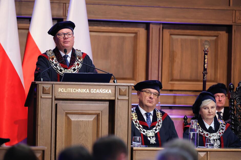 Na zdjęciu rektor w todze i z łańcuchem akademickim przemawia z mównicy oznaczonej napisem „Politechnika Gdańska”. Obok niego siedzą inni przedstawiciele władz uczelni w uroczystych strojach akademickich, a w tle widoczne są biało-czerwone flagi.
