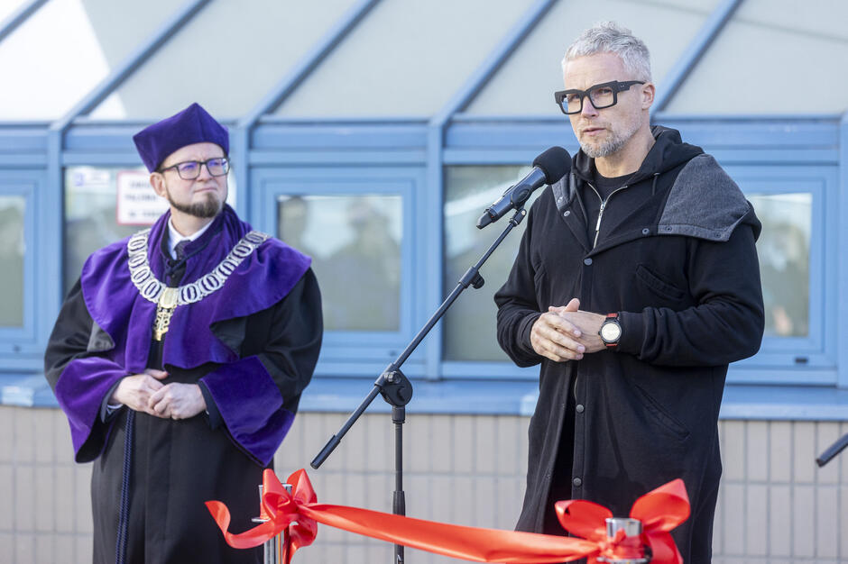 Na zdjęciu widać uroczystość odsłonięcia rzeźby – przy mikrofonie przemawia mężczyzna w czarnym stroju, a obok niego stoi dziekan Wydziału Prawa i Administracji UG w todze z fioletowym kołnierzem i łańcuchem. Przed nimi widoczna jest czerwona wstęga symbolizująca oficjalne otwarcie wydarzenia.

