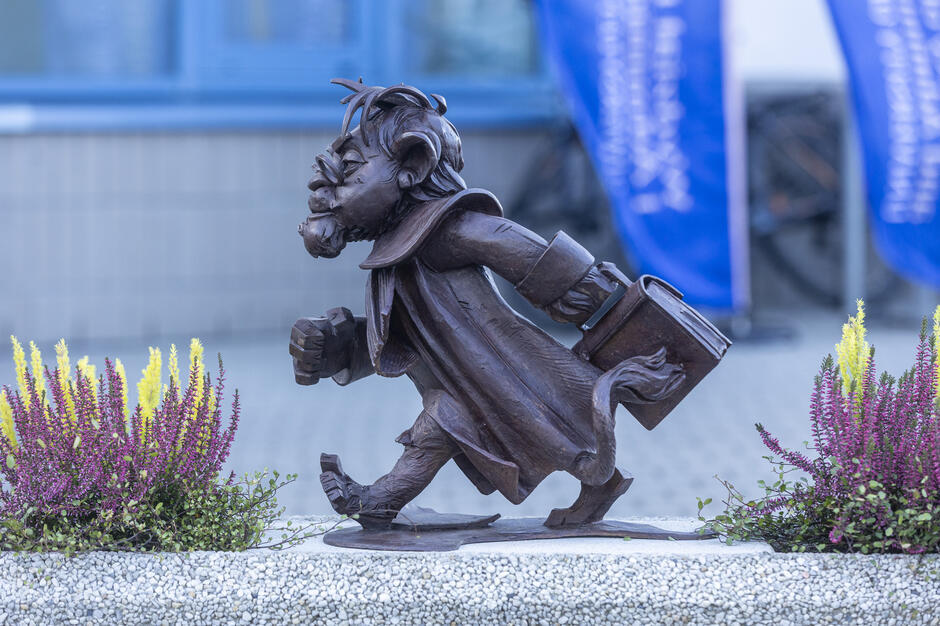 Na zdjęciu znajduje się rzeźba Heweliona przedstawiająca lwa-prawnika w todze, dynamicznie kroczącego z teczką w łapie. Postać symbolizuje energię i zaangażowanie w pracę prawnika, a całość otoczona jest kolorowymi kwiatami.
