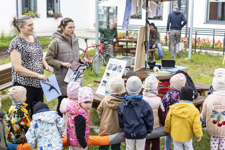 Dwie kobiety na stoisku edukacyjnym w ogrodzie opowiadają dzieciom o ptakach jerzykach, jedna z kobiet trzyma zdjęcie ptaka.
