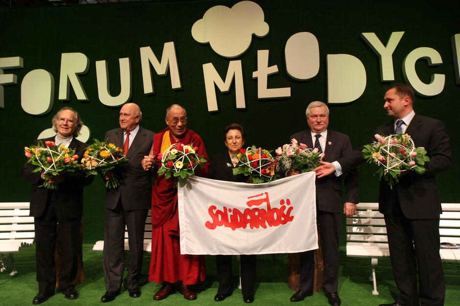 Grupa kilku osób pozuje do zdjęcia na scenie, trzymają flagę z napisem Solidarność .