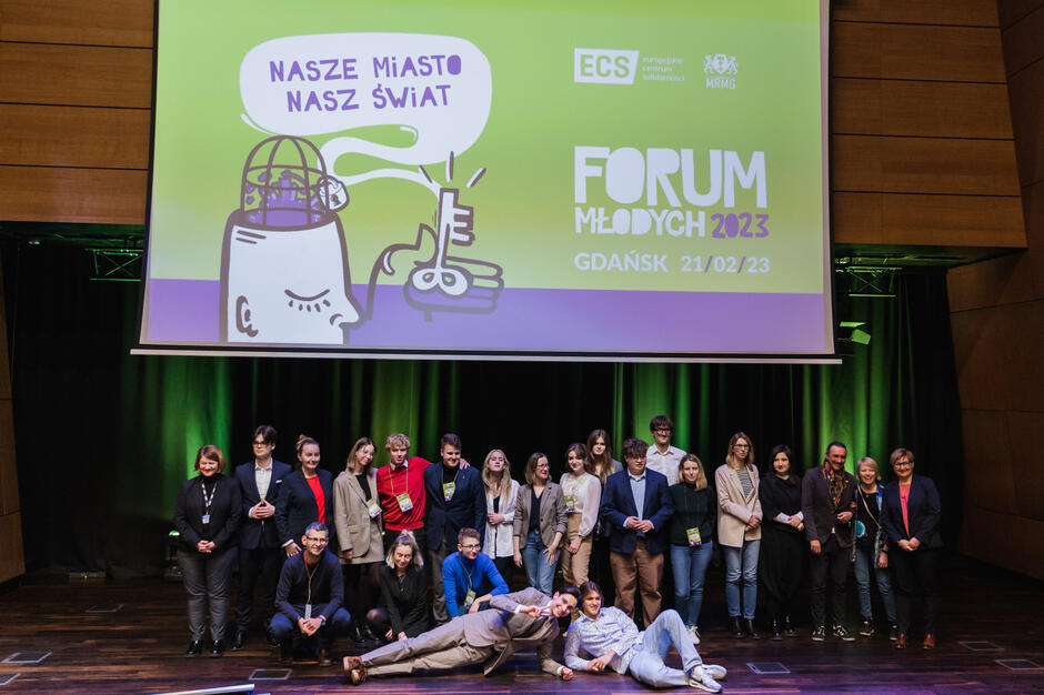 Liczna grupa młodych ludzi pozuje do zdjęcia na scenie, część z nich stoi, część leży. Nad nimi ekran ze slajdem z grafiką głowy i mózgu oraz napisem Forum Młodych .