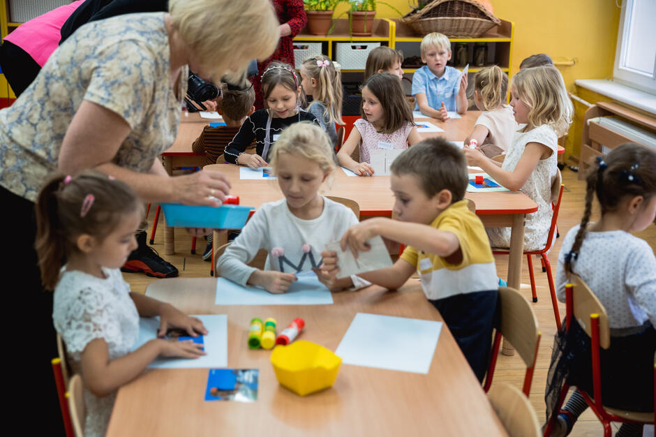 Dzieci siedzą przy stolikach w sali przedszkolnej i wykonują prace plastyczne z kartkami oraz klejem. Nauczycielka pomaga im w trakcie zajęć, podając potrzebne materiały.
