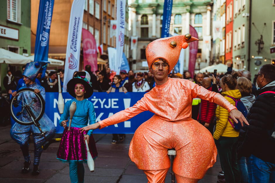 Artyści w kolorowych, fantazyjnych strojach biorą udział w ulicznej paradzie, prezentując jazdę na monocyklu i żonglerkę. Za nimi idzie tłum uczestników oraz widzowie, którzy obserwują wydarzenie w centrum miasta.
