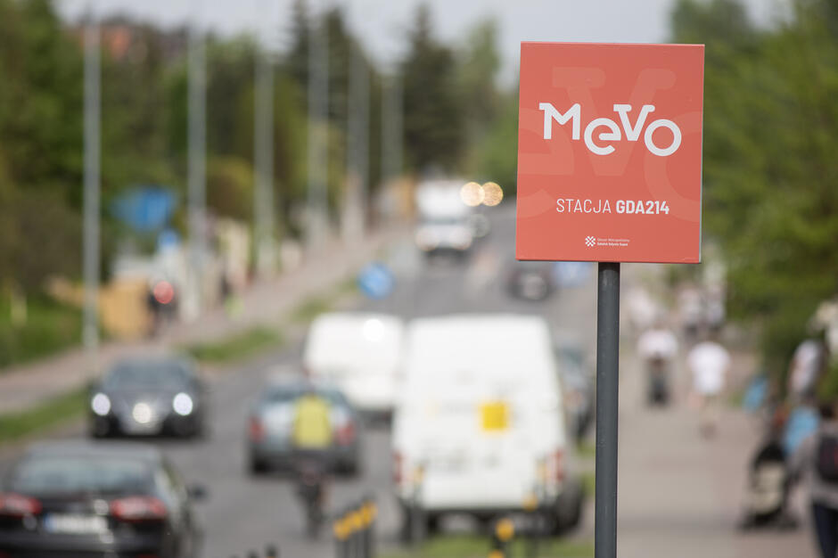 Na pierwszym planie znajduje się czerwony znak systemu rowerów miejskich Mevo z napisem „STACJA GDA214”. W tle widać ruchliwą ulicę z samochodami, rowerzystą oraz pieszymi poruszającymi się chodnikiem.
