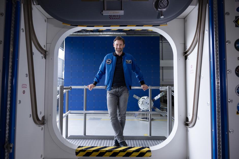 Mężczyzna w niebieskiej kurtce pozuje do zdjęcia uśmiechając się w otoczeniu przypominającym stację kosmiczną.