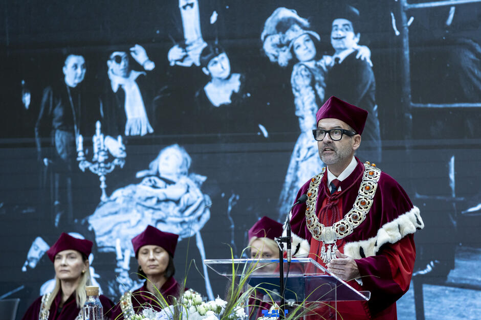 Rektor w uroczystej todze i insygniach przemawia z mównicy podczas inauguracji roku akademickiego. W tle wyświetlona jest czarno-biała scena teatralna, nawiązująca do twórczości Witkacego. 