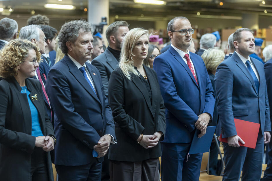 Zaproszeni goście w eleganckich strojach uczestniczą w inauguracji roku akademickiego, stojąc podczas oficjalnej części uroczystości. W ich dłoniach widoczne są teczki z dokumentami, co podkreśla formalny charakter wydarzenia. 