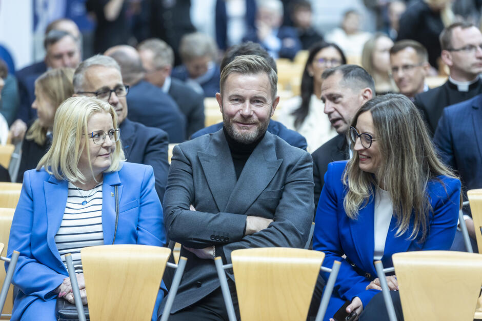 Goście wydarzenia siedzą w auli, prowadząc rozmowy i uśmiechając się do siebie w oczekiwaniu na rozpoczęcie uroczystości. Atmosfera jest swobodna i przyjazna, mimo formalnego charakteru spotkania.
