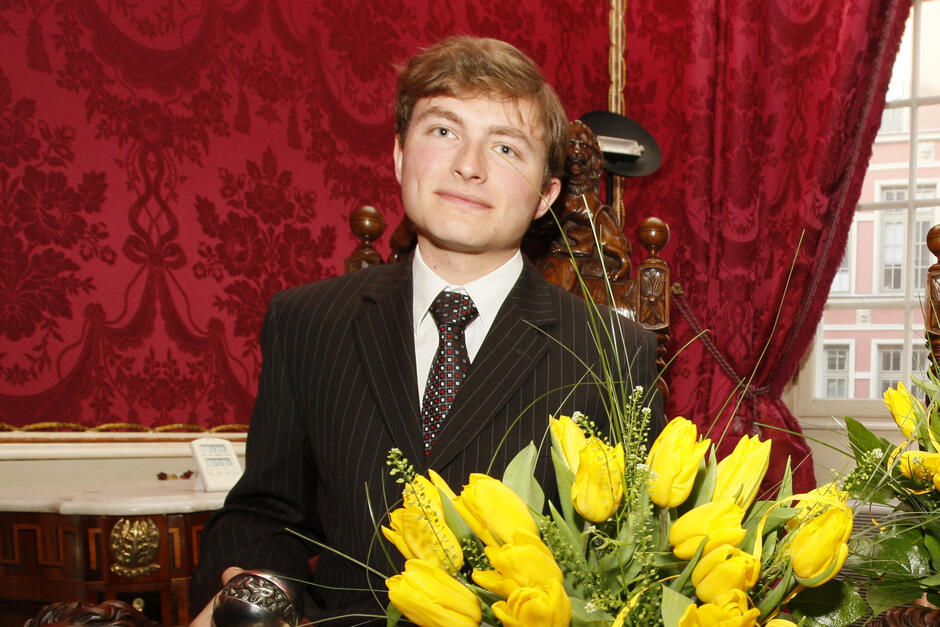 Młody mężczyzna pozuje do zdjęcia w garniturze i z bukietem kwiatów w reprezentacyjnym starym wnętrzu kamienicy.