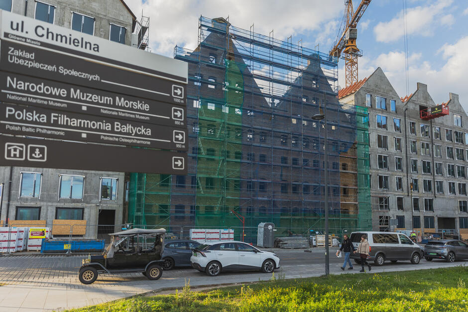 Trwa budowa kompleksu budynków przy ulicy Chmielnej w Gdańsku, gdzie widoczne są rusztowania, żuraw budowlany i nowe fasady z licznymi oknami. Na pierwszym planie znajdują się przechodnie, samochody oraz tablica kierunkowa prowadząca do pobliskich atrakcji, takich jak Narodowe Muzeum Morskie czy Polska Filharmonia Bałtycka. 