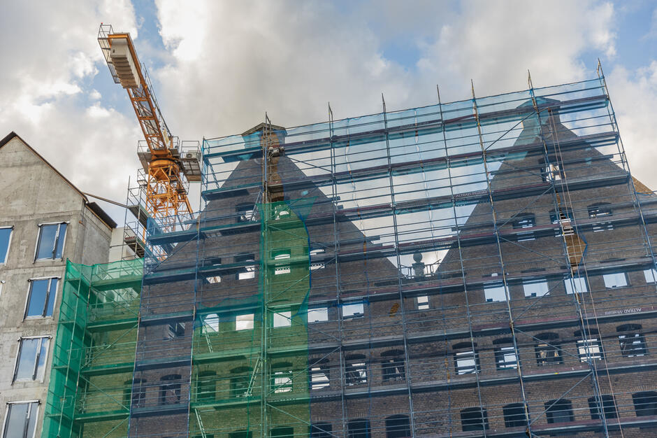 Historyczne spichlerze przykryte są gęstą siatką rusztowań i zabezpieczeń, a nad nimi góruje żuraw budowlany. Na tle nieba wyraźnie widać ceglane szczyty budynków poddawanych renowacji. 