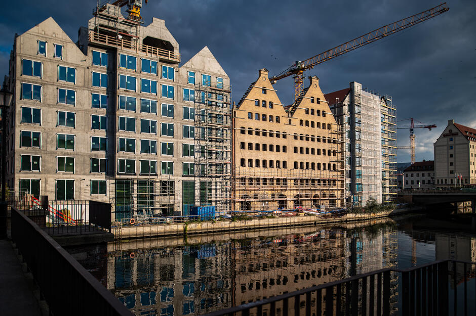 Trwa renowacja i rozbudowa dawnych spichlerzy nad Motławą, otoczonych rusztowaniami i dźwigami budowlanymi. Na rzece widać dwóch kajakarzy płynących obok powstających zabudowań. 