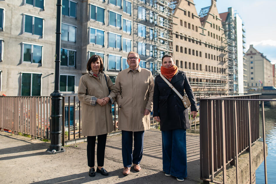 Trzy osoby stoją na nabrzeżu nad Motławą w Gdańsku, pozując do zdjęcia na tle odnawianych spichlerzy i nowych budynków w trakcie budowy. Ubrane są w płaszcze chroniące przed chłodem, a za nimi widoczne są rusztowania i żurawie budowlane. 