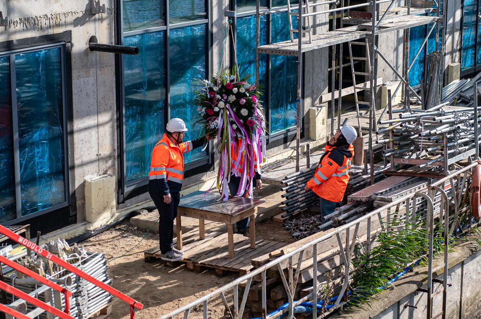 Dwóch pracowników w pomarańczowych kamizelkach i kaskach ustawia na stole wieniec udekorowany kolorowymi wstążkami. Scena odbywa się na placu budowy przy rusztowaniach i materiałach konstrukcyjnych, w tle widoczne są nowe okna budynku.
