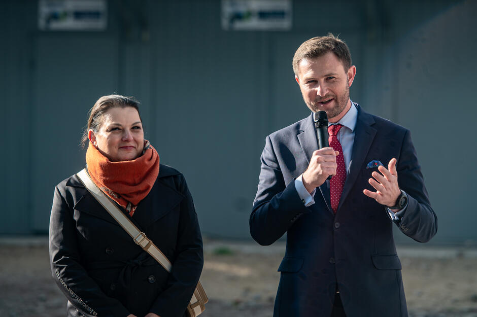 Mężczyzna w garniturze i czerwonym krawacie przemawia do mikrofonu, wykonując gest ręką. Obok niego stoi kobieta w ciemnym płaszczu z pomarańczowym szalikiem, patrząc z uśmiechem w jego stronę.
