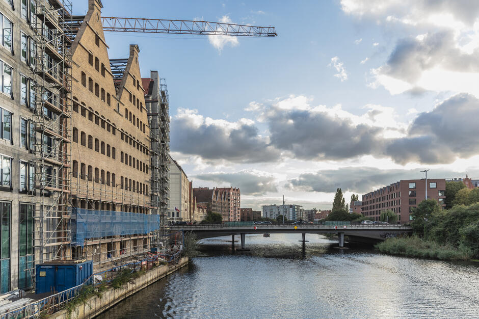Widok na zabudowę Wyspy Spichrzów w Gdańsku, gdzie trwają prace remontowe przy ceglastych spichlerzach otoczonych rusztowaniami. Na pierwszym planie płynie Motława, nad którą rozciąga się most z ruchem samochodowym.
