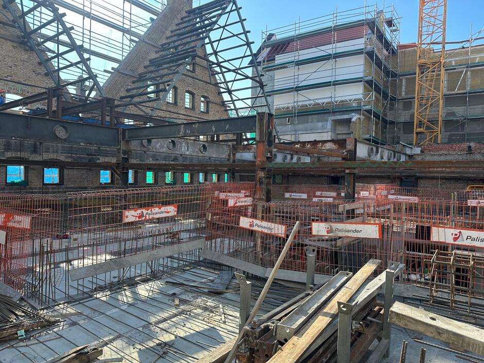 Wnętrze remontowanego spichlerza ukazuje stalowe konstrukcje wsporcze oraz fragmenty zachowanych ceglanych ścian z charakterystycznymi szczytami. Na pierwszym planie widoczne są zbrojenia, szalunki i elementy budowlane przygotowane do dalszych prac.
