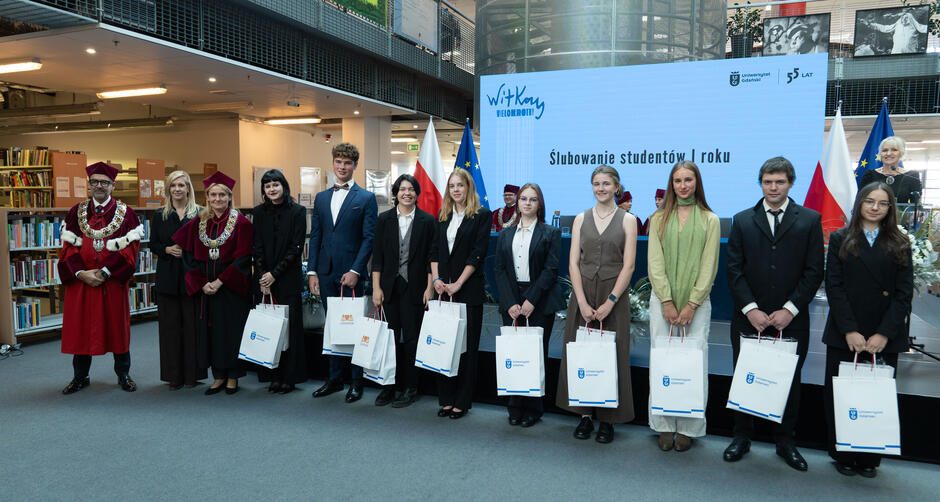 Grupa studentów pierwszego roku Uniwersytetu Gdańskiego stoi wspólnie z władzami uczelni podczas uroczystości ślubowania. Uczestnicy wydarzenia trzymają torby z upominkami, a w tle widoczny jest ekran z napisem „Ślubowanie studentów I roku”. 