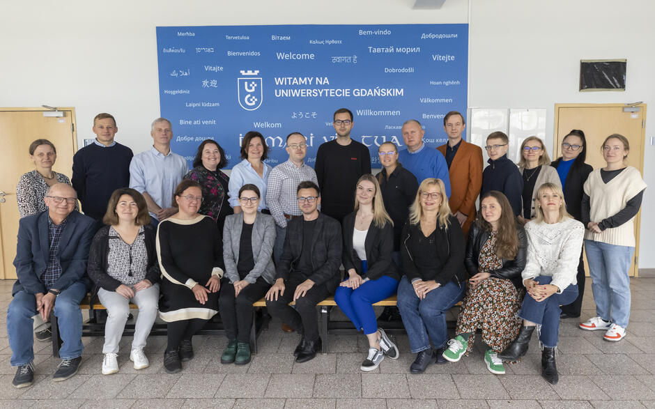 Grupowe zdjęcie pracowników i współpracowników Uniwersytetu Gdańskiego wykonane na tle ściany z napisem „Witamy na Uniwersytecie Gdańskim” w różnych językach. Uczestnicy stoją i siedzą w dwóch rzędach, uśmiechając się do obiektywu. 