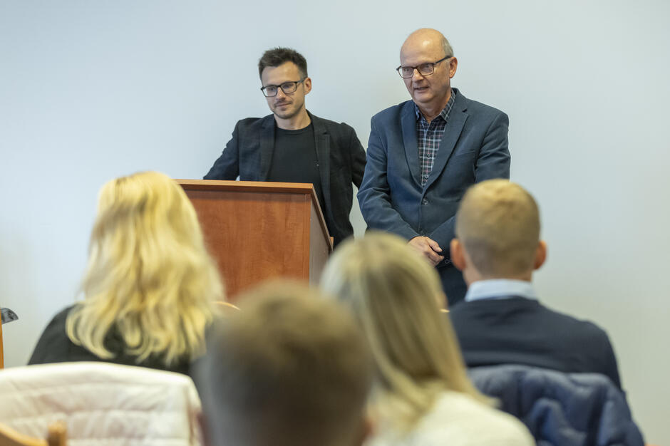 Dwaj mężczyźni stoją przy mównicy, prowadząc wystąpienie przed grupą słuchaczy. Za nimi znajduje się jasna ściana, a uczestnicy siedzą zwróceni w ich stronę.
