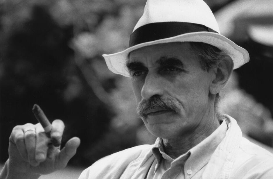 Mężczyzna o szczupłej twarzy, w jasnym kapeluszu, z wąsem. W dłoni trzyma cygaro, które widzimy po lewej