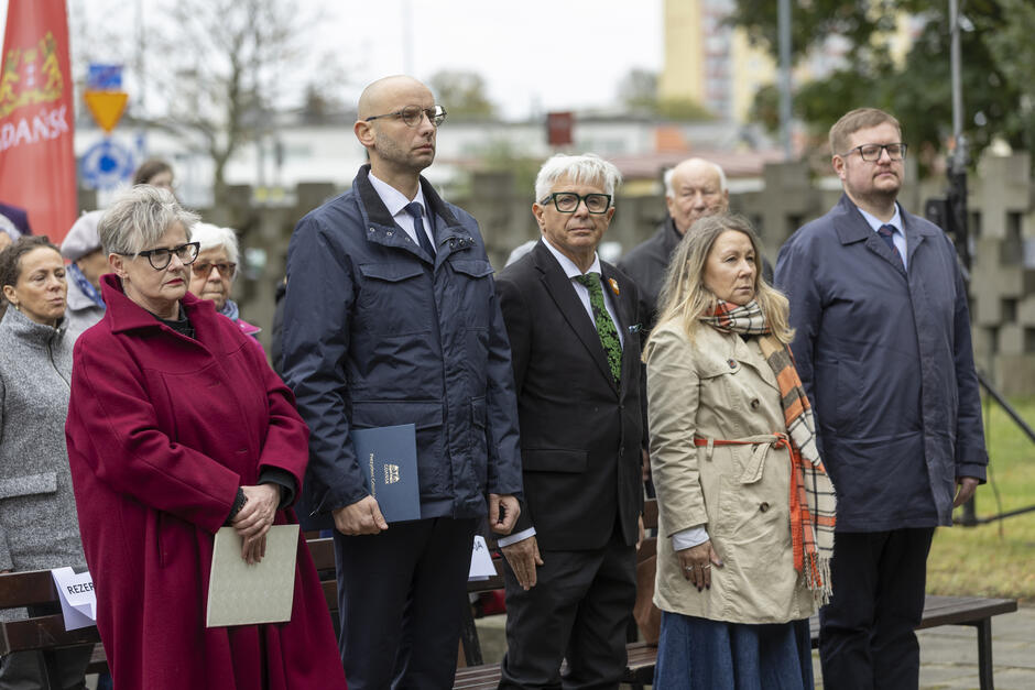 Kilka osób stoi w rzędzie podczas uroczystości na cmentarzu