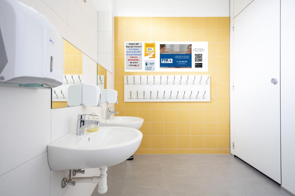Wnętrze jasnej, czystej łazienki z żółtymi płytkami na jednej ścianie i białymi na pozostałych. Po lewej stronie znajdują się dwa białe umywalki z lustrami, mydłem w płynie i automatycznymi podajnikami papieru. Na ścianie naprzeciwko luster wisi wizualizacja tablicy informacyjnej z plakatami dotyczącymi pomocy psychologicznej i wsparcia w sytuacjach kryzysowych. Teksty plakatów są w języku polskim i dotyczą depresji, przemocy, lęku, oraz informują o dostępnych liniach pomocowych i numerach telefonów. Poniżej plakatów wisi rząd wieszaków na ręczniki lub ubrania. Po prawej stronie znajduje się zamknięta kabina WC.