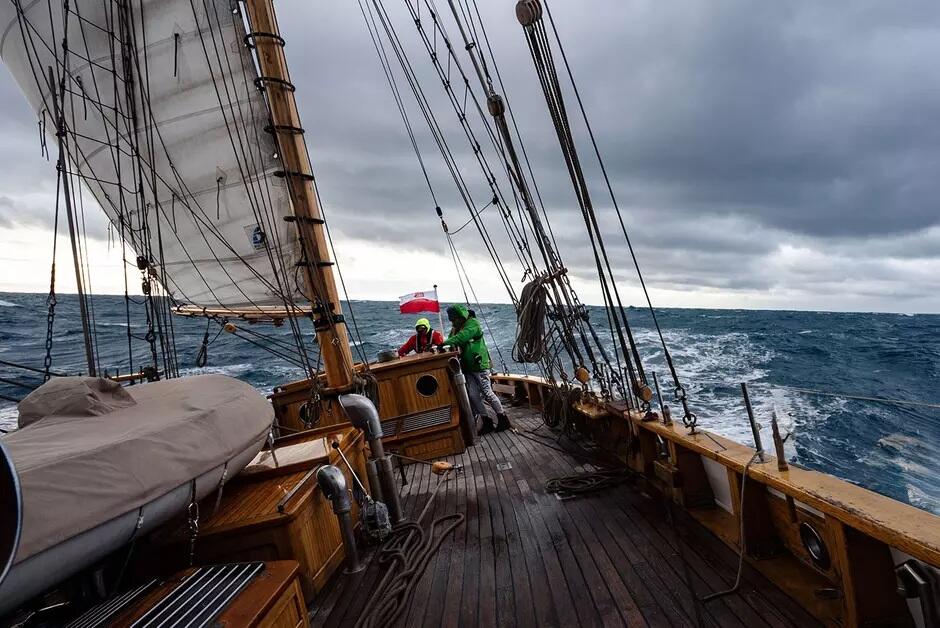 Na zdjęciu żaglowiec płynie po wzburzonym morzu pod ciemnym, burzowym niebem. Dwóch żeglarzy przy sterze, obok powiewającej biało-czerwonej flagi, walczy z wiatrem i falami.
