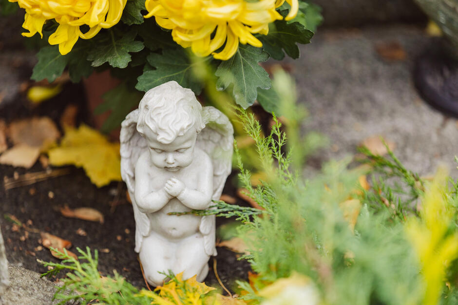 Mała figurka aniołka złożonego do modlitwy stoi wśród zieleni i żółtych kwiatów. Scena ma spokojny, refleksyjny nastrój, przypominający o pamięci i zadumie.
