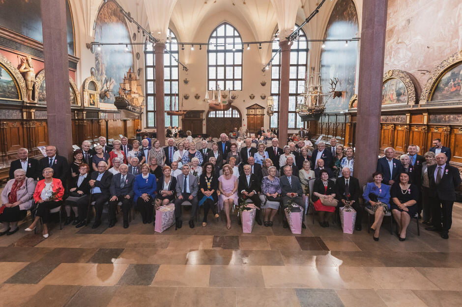 Na zdjęciu duża grupa ludzi zebranych w wielkiej sali z wysokimi sufitami i ozdobnymi dekoracjami. na tylnej ścianie widać duże okna i zwisający z sufitu żyrandol. Kilka kolumn po obu stronach wykonanych jest z marmurowych płytek. Grupa osób jest ustawiona w rzędy, z niektórymi osobami siedzącymi na krzesłach, a innymi stojącymi. Wszyscy są ubrani w formalne stroje, w tym garnitury i krawaty, sugerujące, że biorą udział w formalnym wydarzeniu lub spotkaniu. Niektórzy ludzie trzymają bukiety kwiatów