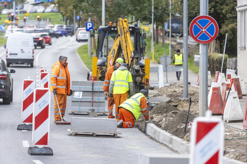 plac budowy na ruchliwej ulicy. Jest czterech robotników budowlanych w pomarańczowych kamizelkach o wysokiej widoczności i kaskach, z boku jest żółta koparka i znak zakazujący zatrzymywania się. na ulicy po lewej stronie zdjęcia jest kilka samochodów 