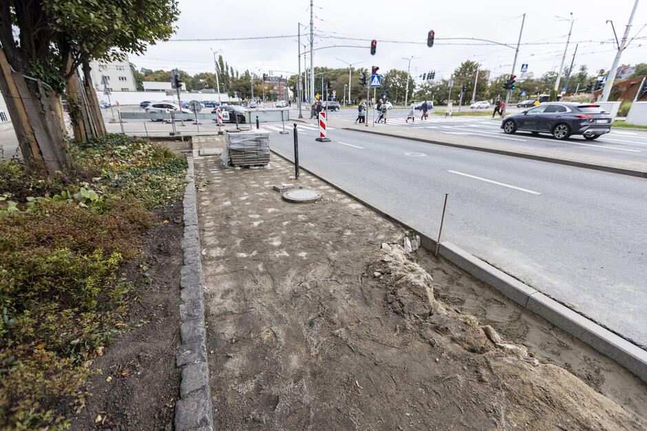 na zdjęciu ślad po rozebranym chodniku, po lewej rosnące duże drzewa zielone, po prawej jezdnia
