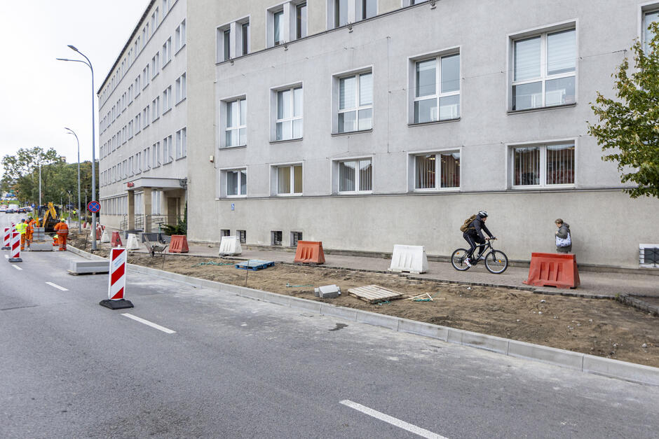 na zdjęciu fragment szarego wysokiego budynku, przed nim rozkopany rozebrany chodnik