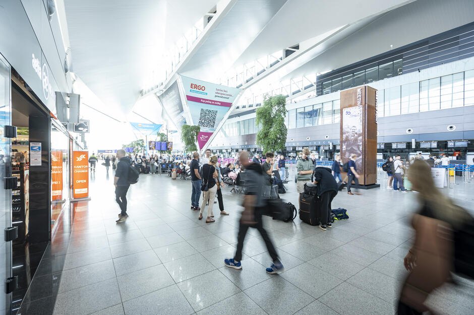 Wnętrze nowoczesnego terminala lotniczego z wysokim, jasnym sufitem i dużą przestrzenią dla podróżnych. Widać kolejki do odprawy bagażowej, ludzi z walizkami oraz sklepy i punkty usługowe po bokach.
