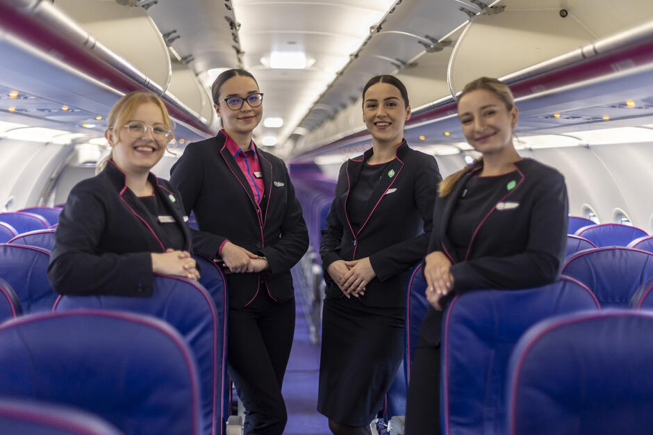 Cztery stewardesy w uniformach WizzAir stoją w przejściu samolotu, uśmiechając się do obiektywu. Wnętrze maszyny wypełniają rzędy niebieskich foteli z różowymi obszyciami. 