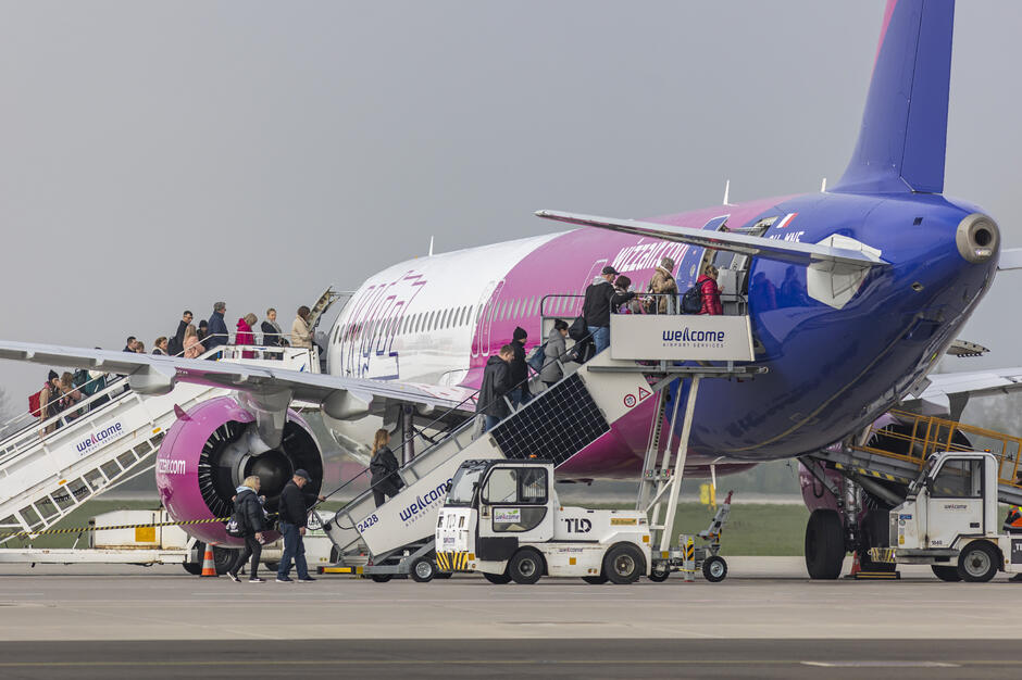 Pasażerowie wchodzą na pokład samolotu WizzAir podstawionego na płycie lotniska. Obsługa naziemna nadzoruje boarding przy pomocy mobilnych schodów i pojazdów serwisowych. 