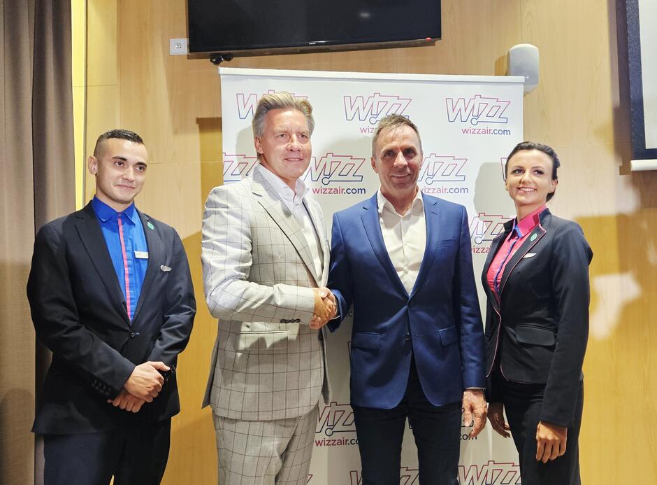 Zdjęcie przedstawia cztery osoby w eleganckich strojach podczas oficjalnego wydarzenia związanego z liniami lotniczymi Wizz Air. Dwaj mężczyźni w centrum uściskiem dłoni potwierdzają współpracę, a w tle widoczne jest logo Wizz Air.
