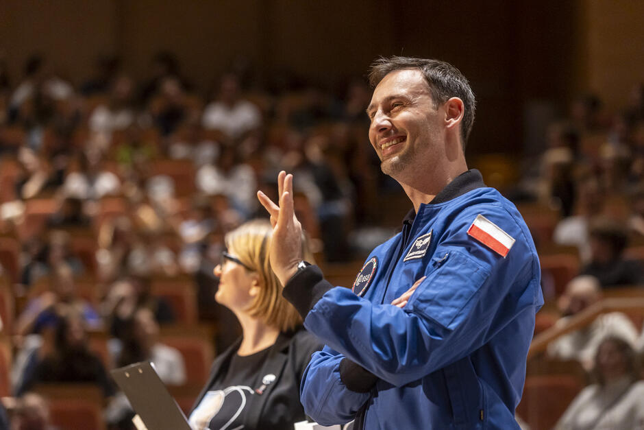 Mężczyzna w kurtce astronauty śmieje się na scenie podczas spotkania z dziećmi.