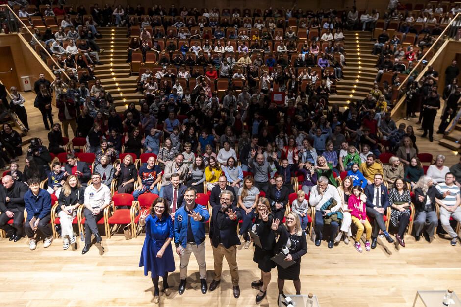 Trzy kobiety i dwóch mężczyzn stoi na scenie i patrzy się w górę do fotografa, wszyscy się śmieją, za nimi widownia - dorośli i dzieci.