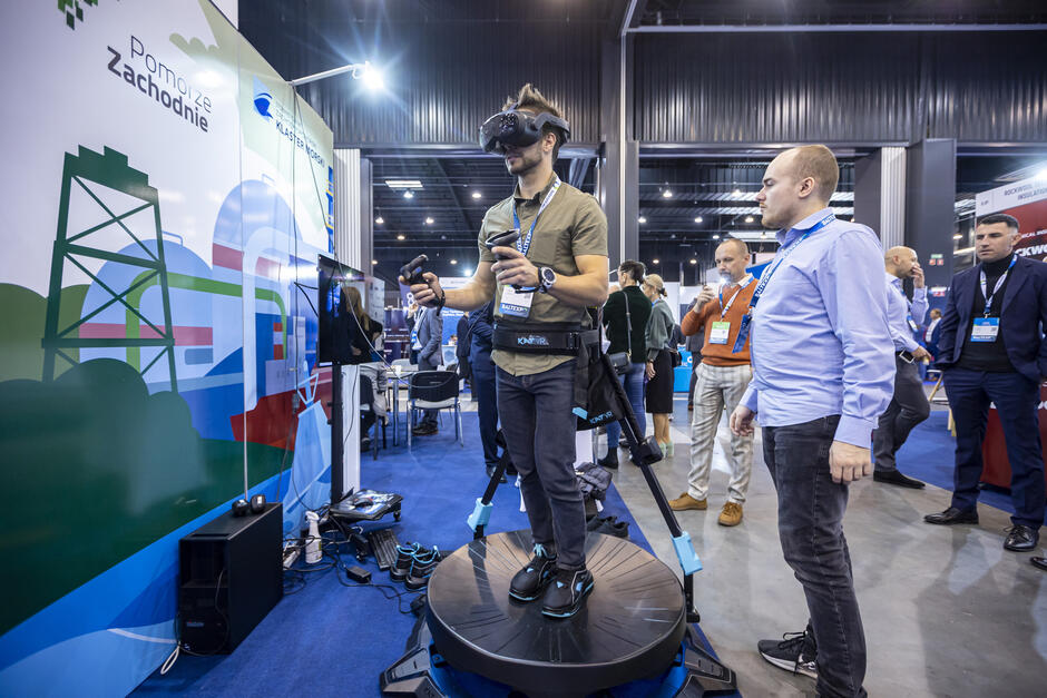 Na zdjęciu widać uczestników targów lub konferencji technologicznej. Mężczyzna w centrum stoi na platformie do wirtualnej rzeczywistości (VR), wyposażony w okulary VR, kontrolery i specjalne obuwie umożliwiające poruszanie się w symulowanym świecie. Obok niego stoi inny uczestnik obserwujący prezentację. W tle widoczne są stoiska wystawców, banery i osoby rozmawiające. Na ścianie po lewej stronie znajduje się grafika z napisem „Pomorze Zachodnie” oraz logo „Klaster ICT Pomorze Zachodnie”. Całość ma charakter demonstracji nowoczesnych technologii immersyjnych podczas wydarzenia branżowego.