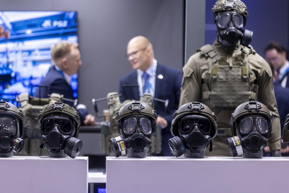 Na zdjęciu zaprezentowano ekspozycję wojskowego sprzętu ochronnego. Na pierwszym planie ustawiono kilka hełmów z zamontowanymi maskami przeciwgazowymi, wyposażonymi w filtry i przyłbice ochronne. W tle stoi manekin ubrany w pełny strój taktyczny z kamizelką balistyczną i maską gazową. Za ekspozycją widoczni są dwaj mężczyźni w garniturach prowadzący rozmowę przy stoisku wystawienniczym. Cała scena odbywa się w profesjonalnym otoczeniu targów lub wystawy obronnej, gdzie prezentowany jest nowoczesny sprzęt dla służb mundurowych.