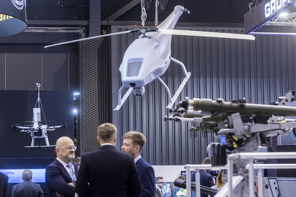 Na zdjęciu znajduje się stoisko wystawowe prezentujące nowoczesne technologie obronne. W centralnej części kadru zawieszony jest duży bezzałogowy śmigłowiec obserwacyjny, wyposażony w kamerę pod kadłubem. W tle widać także mniejszego drona wielowirnikowego zawieszonego na ekspozycji ściennej. Na pierwszym planie po prawej stronie znajduje się element uzbrojenia zamontowany na stanowisku pokazowym. W dolnej części zdjęcia kilku mężczyzn w garniturach prowadzi rozmowę biznesową, co sugeruje charakter targów branżowych poświęconych technologii wojskowej i obronnej.