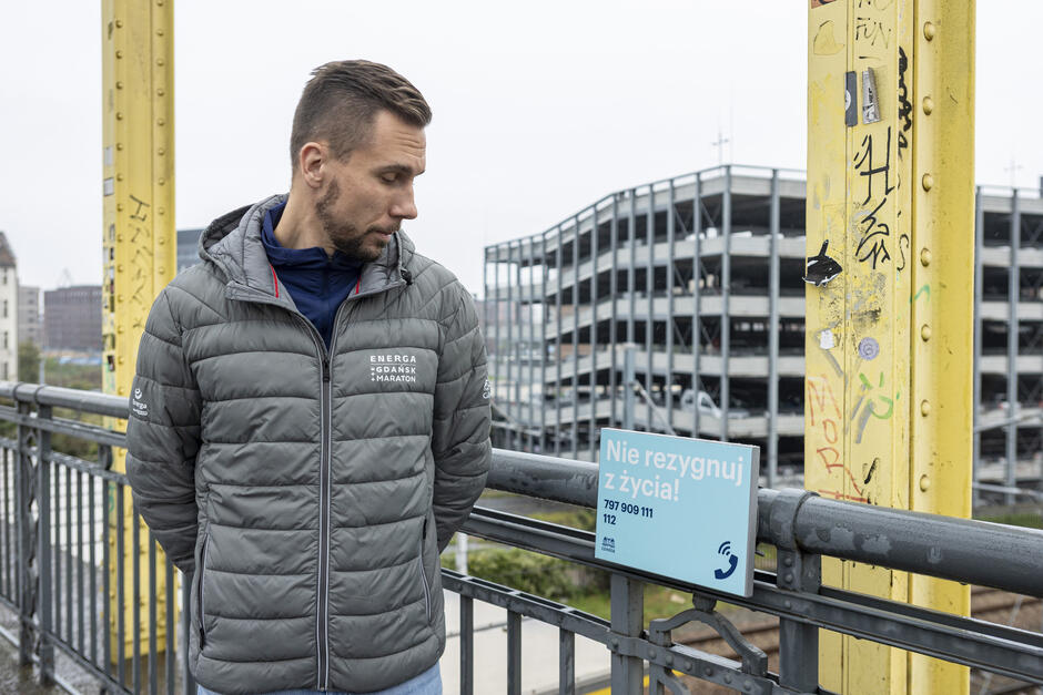 Mężczyzna stoi na wiadukcie i patrzy na tabliczkę z napisem Nie rezygnuj z życia .