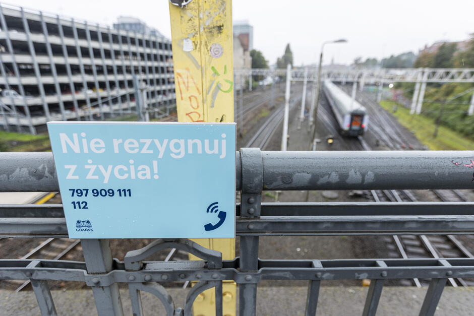 Błękitna tabliczka na wiadukcie z napisem Nie rezygnuj z życia  i numerami telefonów.
