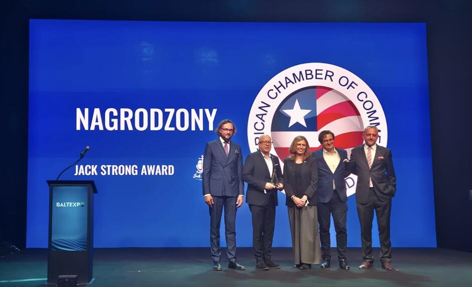 Na zdjęciu widać uroczystość wręczenia nagrody Jack Strong Award podczas wydarzenia Baltexpo. Na scenie, przed dużym ekranem z logotypem American Chamber of Commerce in Poland, stoi pięć osób w eleganckich strojach biznesowych. Jedna z nich trzyma statuetkę symbolizującą otrzymane wyróżnienie. Po lewej stronie znajduje się mównica z logo Baltexpo, a w tle widoczny jest napis „NAGRODZONY – JACK STRONG AWARD”. Scena jest oświetlona profesjonalnym światłem, a atmosfera fotografii ma charakter oficjalny i uroczysty.