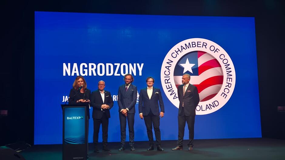 Na zdjęciu widać uroczystość wręczenia nagrody Jack Strong Award podczas wydarzenia Baltexpo. Na scenie, przed dużym ekranem z logotypem American Chamber of Commerce in Poland, stoi pięć osób w eleganckich strojach biznesowych. Jedna z nich trzyma statuetkę symbolizującą otrzymane wyróżnienie. Po lewej stronie znajduje się mównica z logo Baltexpo, a w tle widoczny jest napis „NAGRODZONY – JACK STRONG AWARD”. Scena jest oświetlona profesjonalnym światłem, a atmosfera fotografii ma charakter oficjalny i uroczysty.