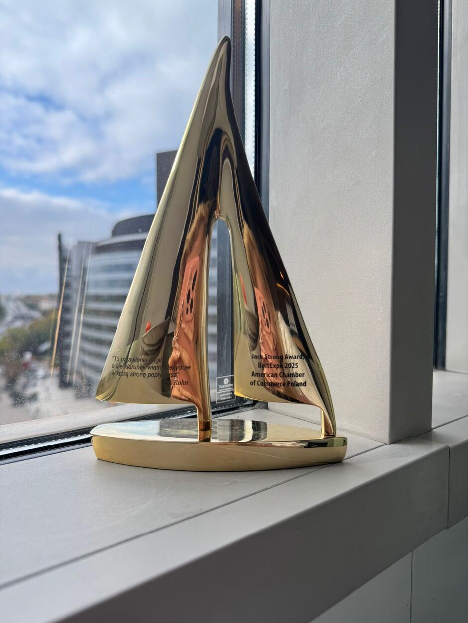 Na zdjęciu przedstawiono statuetkę w kształcie stylizowanego żagla, wykonaną z błyszczącego, złotego metalu. Na powierzchni widoczny jest grawer z napisem „Jack Strong Award Baltexpo 2025 American Chamber of Commerce Poland” oraz cytatem inspirowanym motywem żeglarskim. Nagroda stoi na parapecie przy oknie, w tle widać fragment nowoczesnych budynków i niebo z lekkim zachmurzeniem. Całość kompozycji podkreśla elegancję i prestiż wyróżnienia.